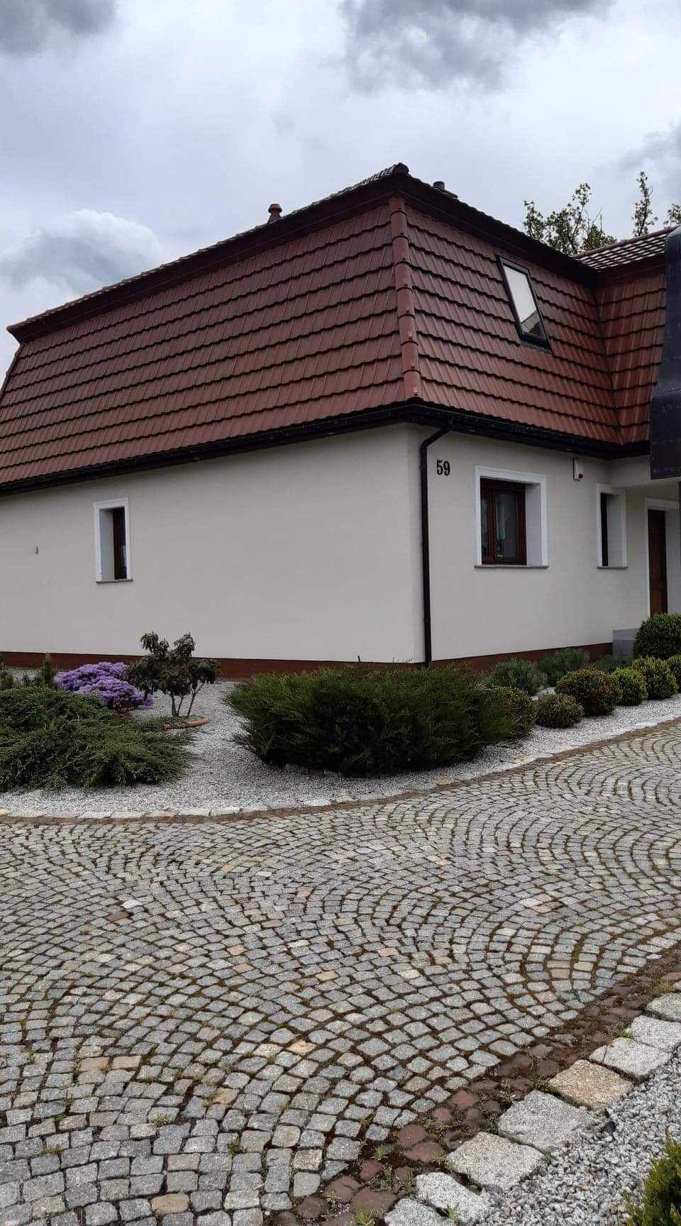 Dom jednorodzinny z brązowym dachem z dachówki, białymi ścianami, numerem 59, małymi oknami z brązowymi ramami i brukowanym podjazdem z kamiennej kostki.