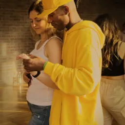 Instruktor w żółtej bluzie i czapce prowadzi kobietę w tańcu, trzymając ją za ręce w jasnym pomieszczeniu z ceglaną ścianą.