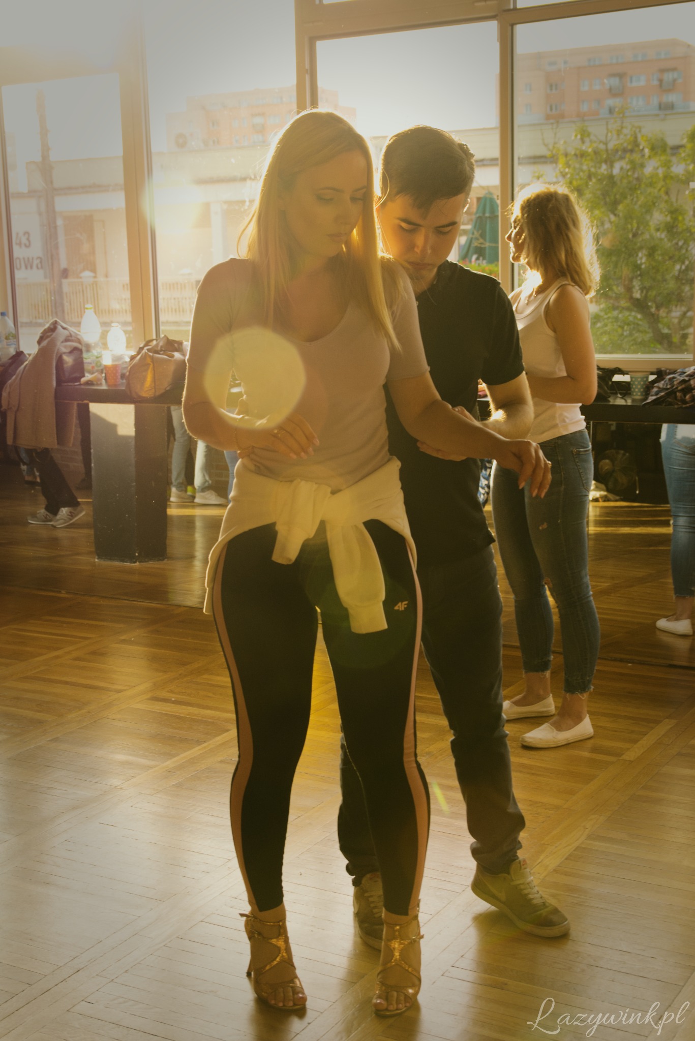 Instruktor tańca poprawia postawę kursantki podczas zajęć w jasnej sali z parkietem, w tle widoczne okna i inne osoby.