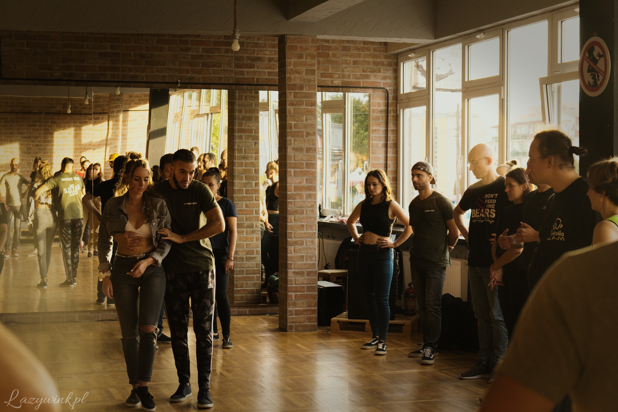 Grupa osób w sali z lustrami, para w centrum w trakcie lekcji, instruktor trzyma ręce na talii kursantki, widoczne ceglane ściany i duże okna w tle.