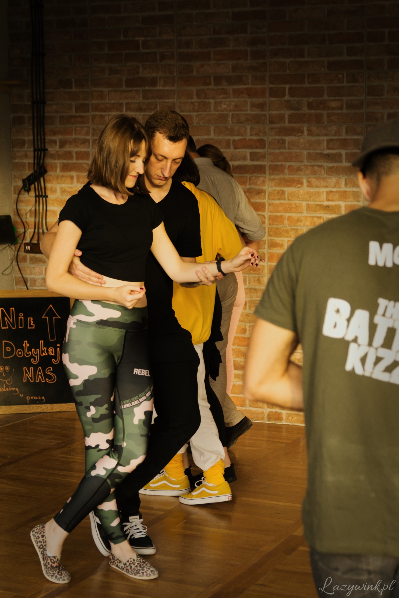 Para taneczna w trakcie zajęć, ona w legginsach moro i baletkach w panterkę, on w czarno-żółtej bluzie i żółtych vansach, w tle ceglana ściana i tablica z napisem 'Nie Dotykaj NAS'.