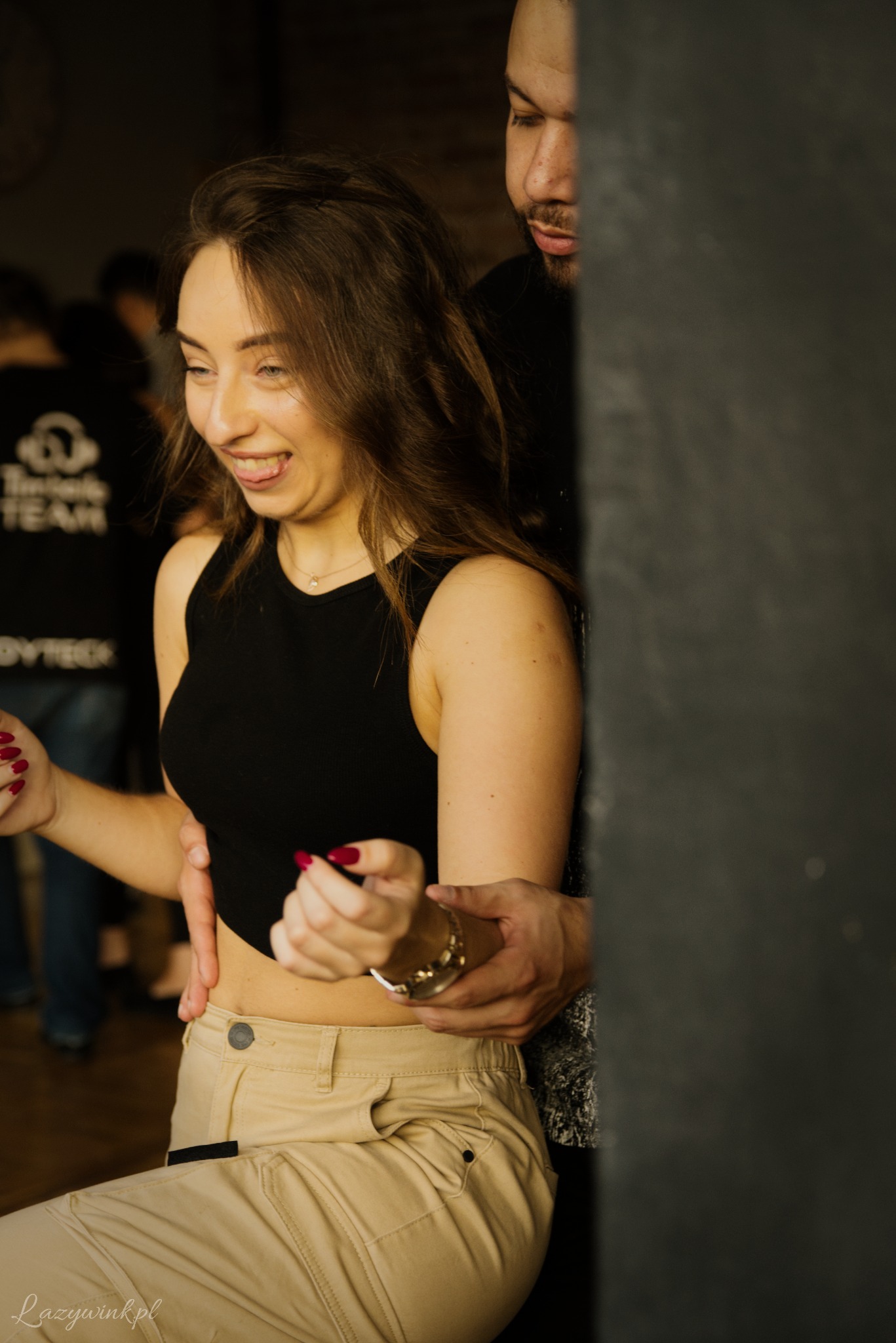 Uśmiechnięta kobieta w czarnym topie i beżowych spodniach, z wyciągniętym językiem, trzymająca bransoletkę, z mężczyzną w tle, w sali z innymi osobami.