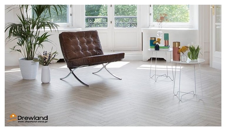 Jasna podłoga drewniana ułożona w jodełkę francuską w salonie z brązowym skórzanym fotelem Barcelona, roślinami w doniczkach i białymi stolikami pomocniczymi, widoczne okna i drzwi balkonowe.