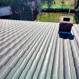 Szary dach z blachy trapezowej z kominem z czerwonej cegły, widok z góry na tle zielonego ogrodu.