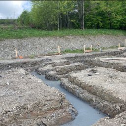 TegesBud - Wykopy pod fundamenty wypełnione wodą. Widoczne drewniane znaczniki i niwelator. W tle zieleń. Prace ziemne przygotowawcze pod budowę.