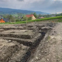 Przygotowany wykop pod fundamenty z widocznym zbrojeniem, w tle zabudowa jednorodzinna i zalesione wzgórza. Prace ziemne na działce budowlanej.