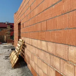 Wznoszenie ściany z czerwonej ceramiki poryzowanej, widoczna paleta oparta o ścianę, fragment budynku w tle.