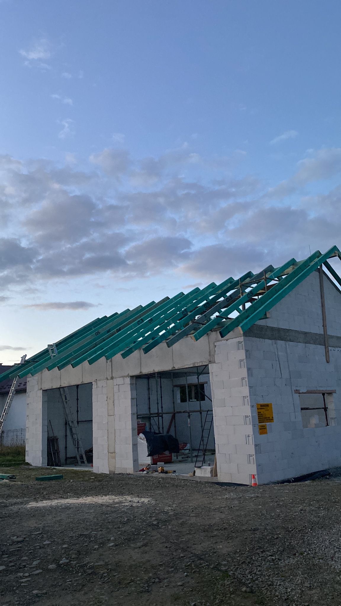 Surowy budynek z bloczków, z zielonymi elementami więźby dachowej, na tle pochmurnego nieba. Widoczne rusztowania i drabina. Budowa w toku.