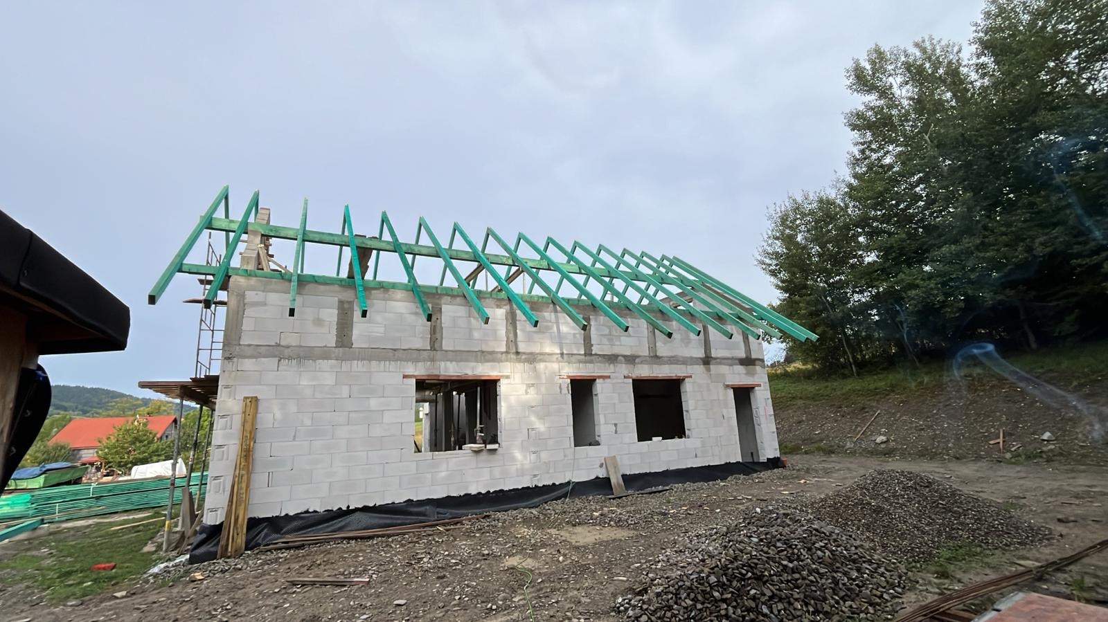 Stan surowy otwarty domu z bloczków w Nowym Sączu, z zieloną konstrukcją dachu. Wokół gruz i teren przygotowany pod dalsze prace.