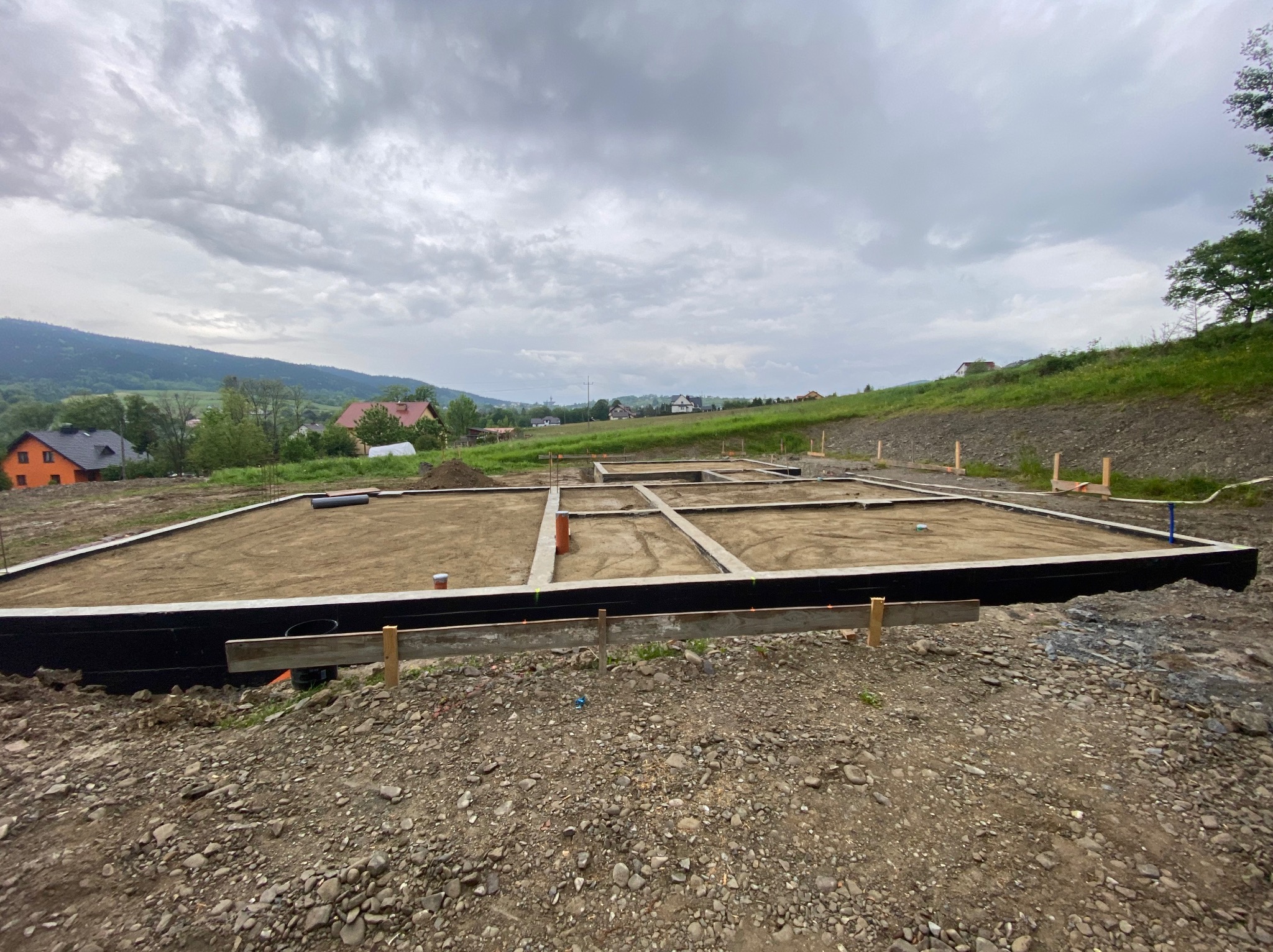 Fundamenty domu jednorodzinnego w trakcie budowy, widok z góry na zbrojenie i szalunki, w tle zielone wzgórza i domy. Pochmurne niebo.