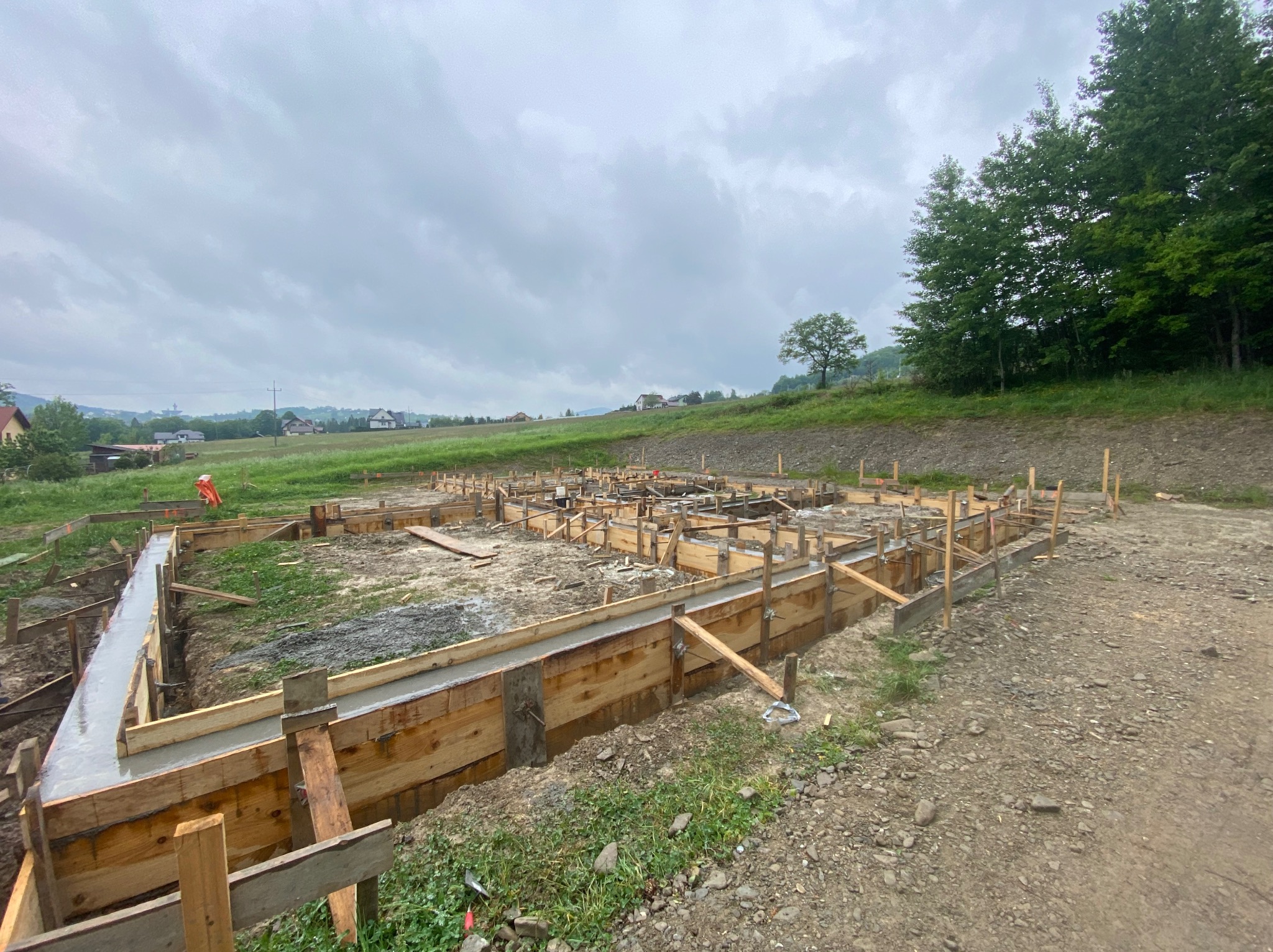 Zalane betonem szalunki fundamentów domu jednorodzinnego na nierównym terenie, widok na zielone wzgórza i pochmurne niebo.