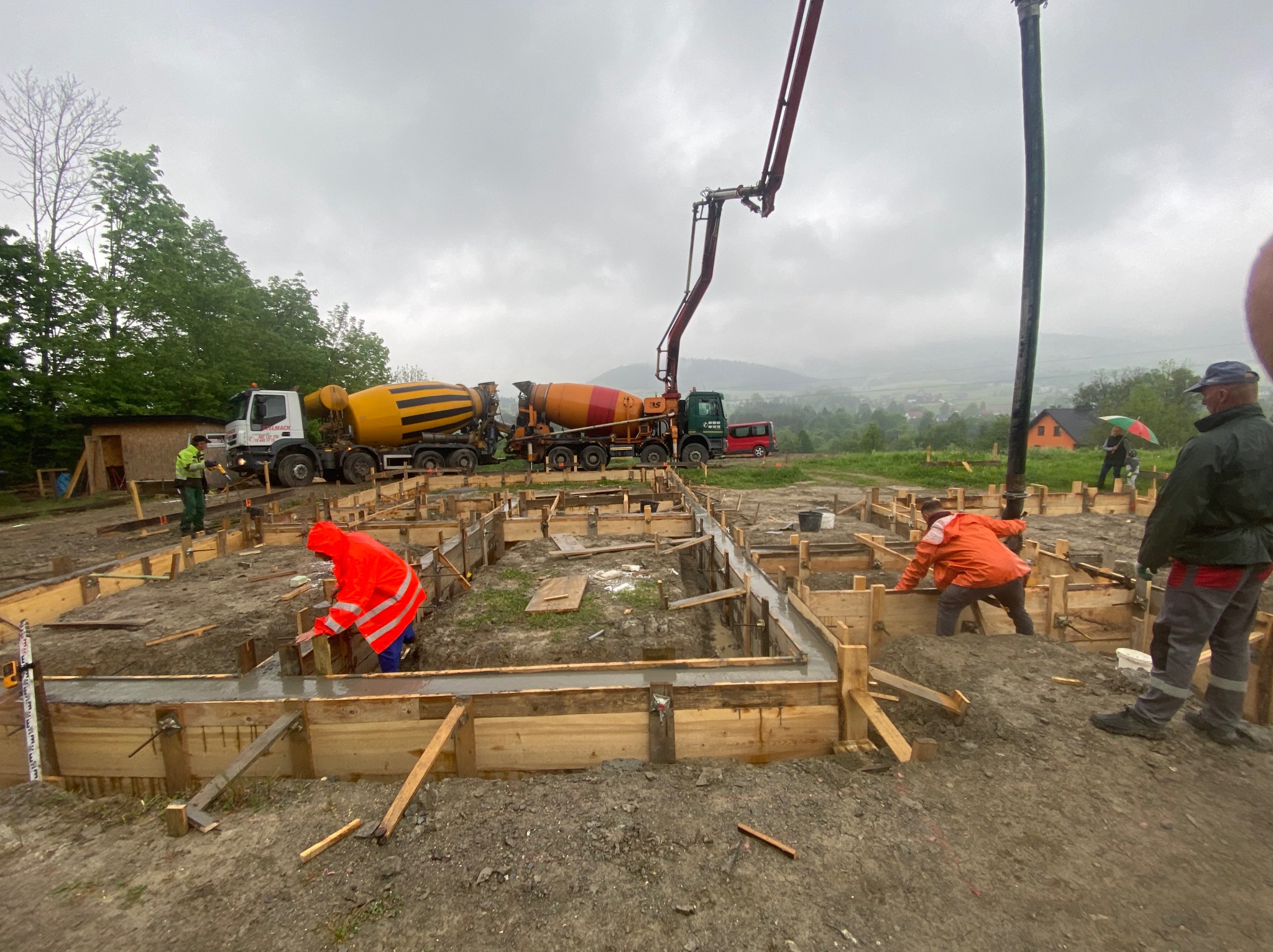Wylewanie fundamentów domu jednorodzinnego. Pracownicy w odblaskowych ubraniach, betonowozy, drewniane szalunki. Krajobraz górski w tle.