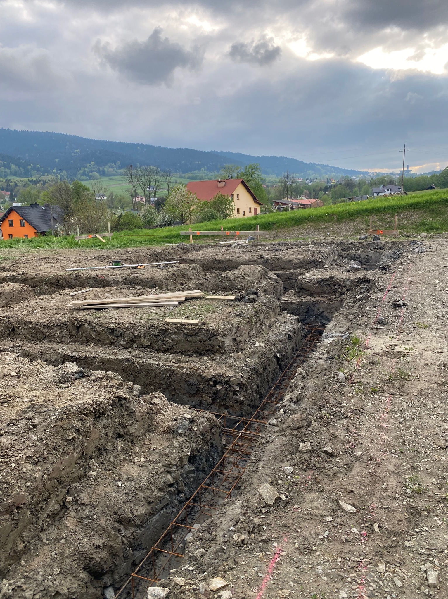 Przygotowany wykop pod fundamenty z widocznym zbrojeniem, w tle zabudowa jednorodzinna i zalesione wzgórza. Prace ziemne na działce budowlanej.