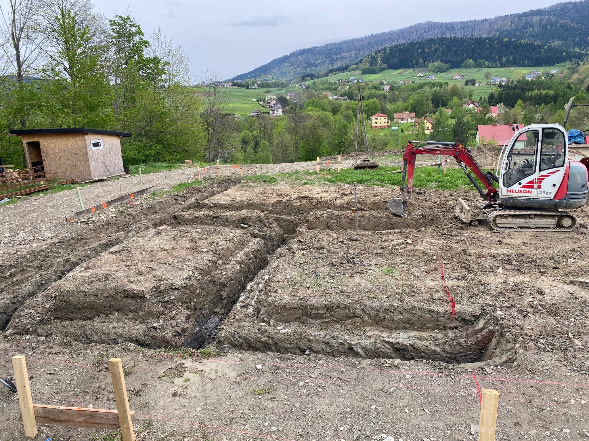 Mini koparka Neuson 2503 przy wykopie fundamentów, widoczne drewniane szalunki i mały barak na tle zielonych wzgórz w pochmurny dzień.