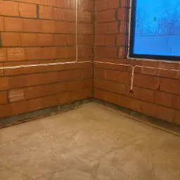 Surowy pokój z czerwonej cegły z poprowadzonymi przewodami elektrycznymi w białych listwach, widoczne puszki instalacyjne i okno z widokiem na zimowy krajobraz.