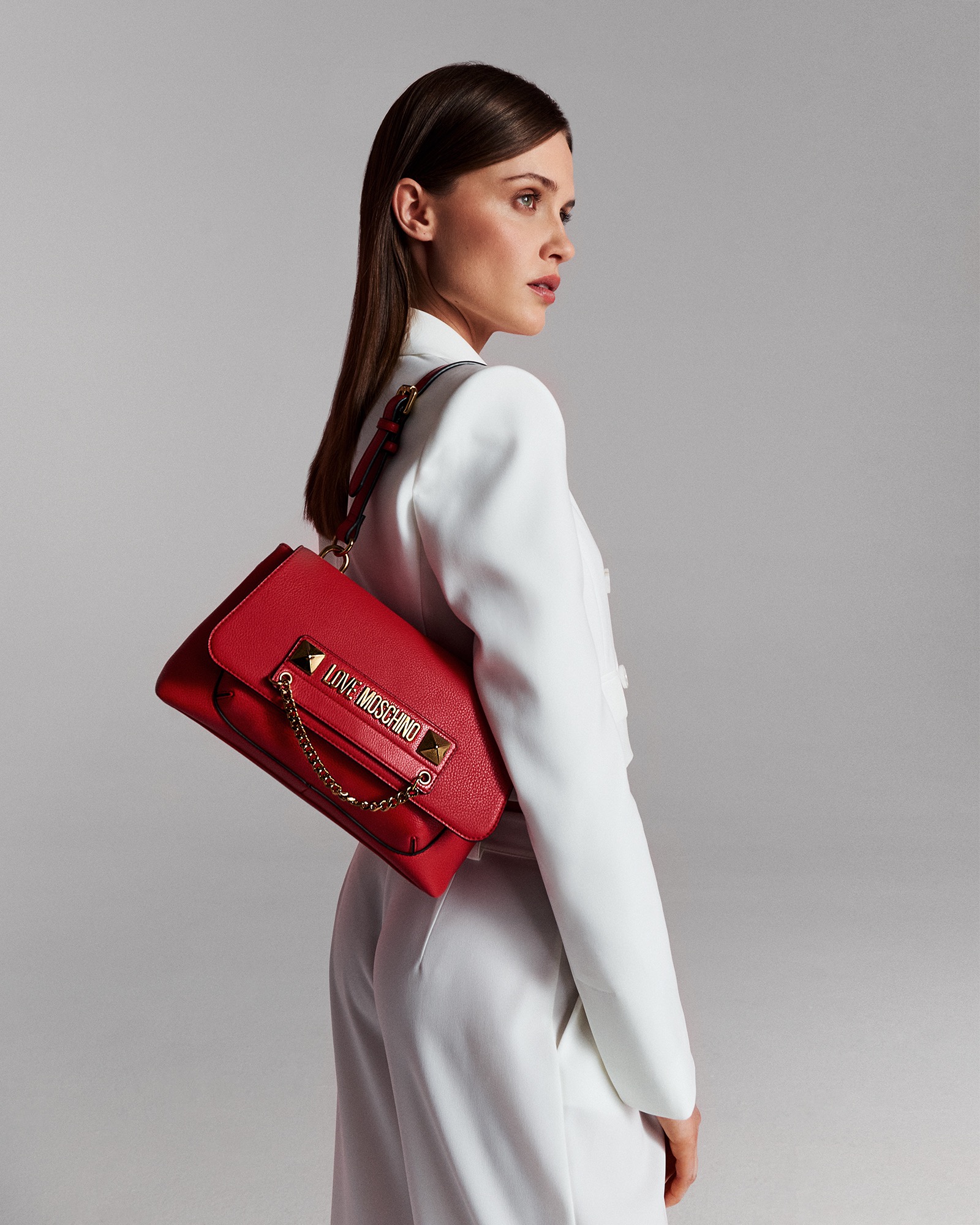 Kobieta w białym garniturze prezentuje czerwoną torebkę marki Love Moschino z łańcuszkiem i złotymi detalami