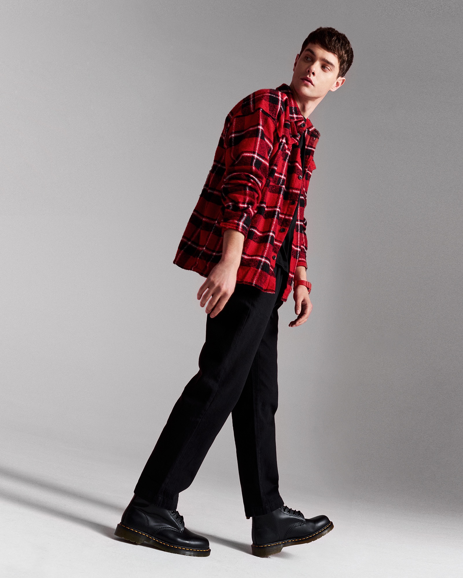 Mężczyzna w czerwonej koszuli w kratę, czarnych spodniach i butach marki Dr. Martens, pozujący na jasnoszarym tle, z profilu.