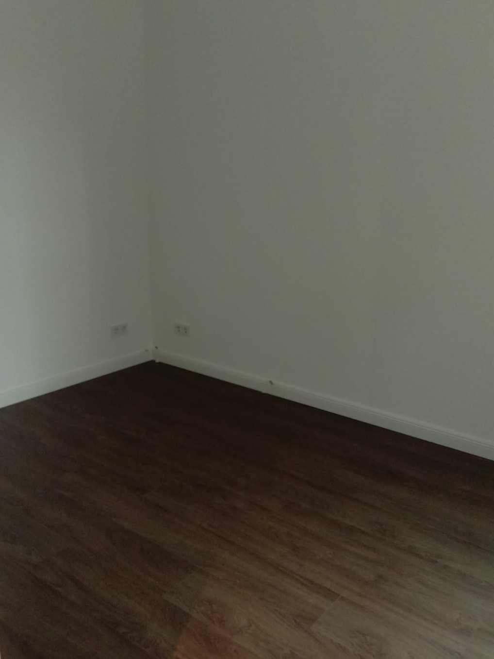 Kąt pokoju z brązową podłogą imitującą drewno, białe ściany z listwami przypodłogowymi i gniazdkami elektrycznymi.