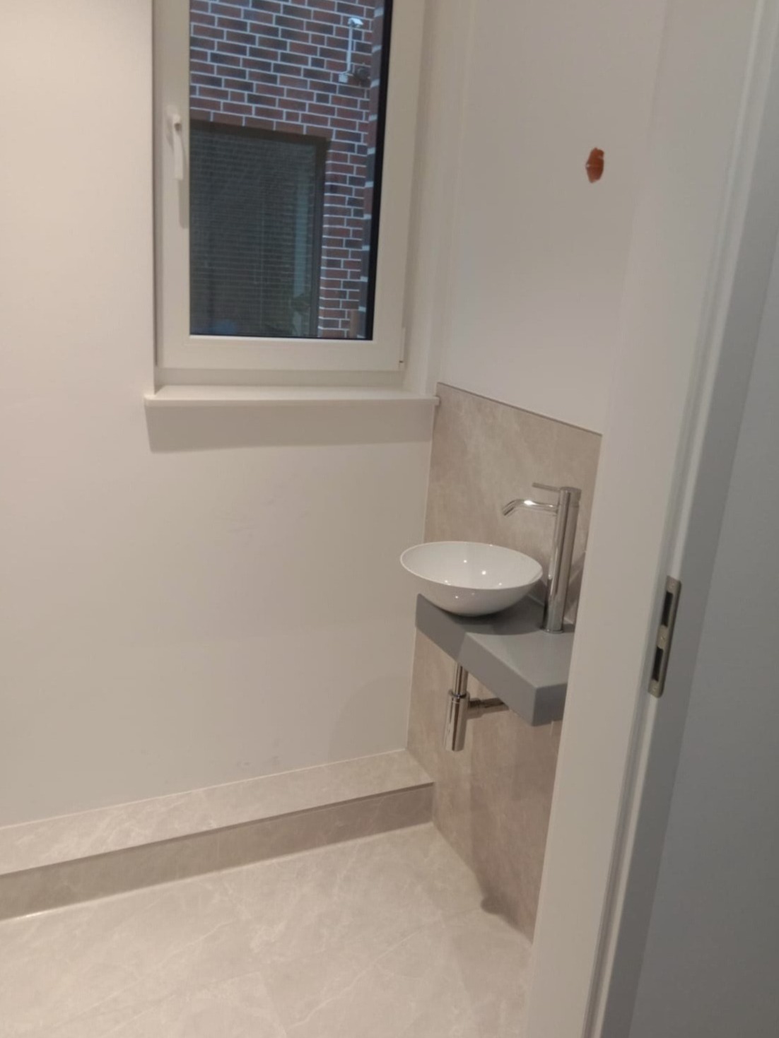 Nowoczesna, minimalistyczna łazienka z umywalką nablatową na szarym blacie, chromowaną baterią i beżowymi płytkami imitującymi marmur, widok na okno z ceglanym budynkiem na zewnątrz.