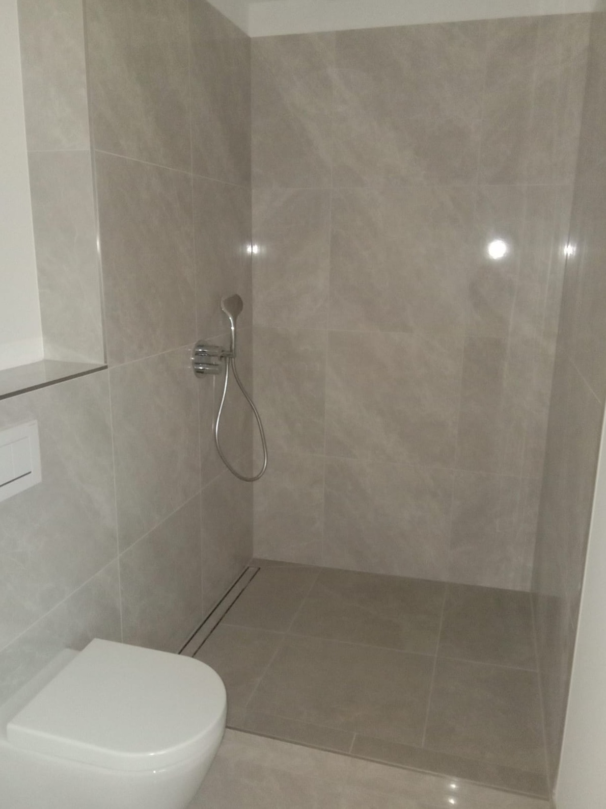 Nowoczesna kabina prysznicowa z odpływem liniowym i armaturą w kolorze chromu, wykończona płytkami imitującymi marmur, z widoczną częścią toalety.