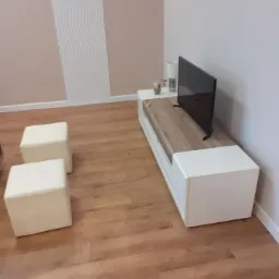 Jasne panele podłogowe w salonie, biała szafka RTV z drewnianym blatem, telewizor, dwie białe pufy i dekoracyjna biała listwa na ścianie w tle.