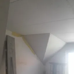 Poddasze w trakcie ocieplania, widoczne płyty gipsowo-kartonowe na skosach, zabezpieczone taśmą łączenia, drewniane deski na podłodze, rusztowanie przy oknie.