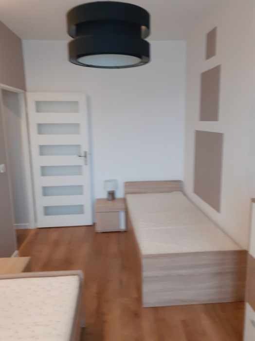 Wnętrze pokoju po odświeżeniu: drewniana podłoga, pojedyncze łóżko z szafką nocną, białe drzwi z przeszkleniami i nowoczesna lampa sufitowa.