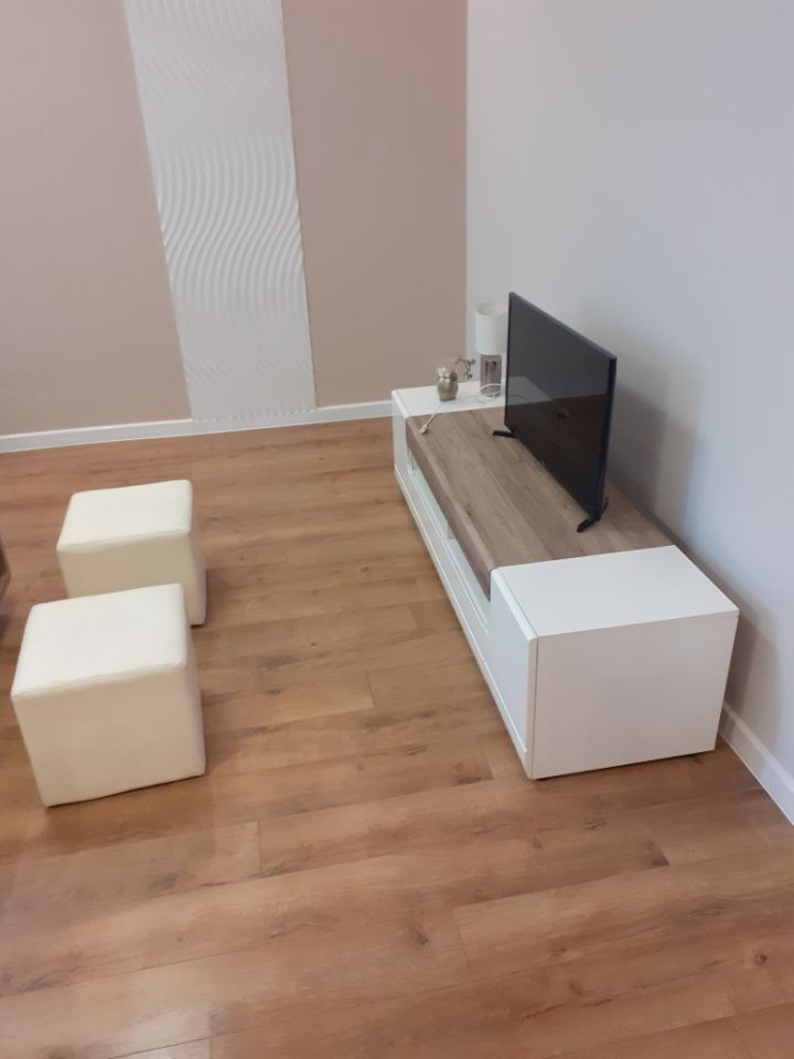 Jasne panele podłogowe w salonie, biała szafka RTV z drewnianym blatem, telewizor, dwie białe pufy i dekoracyjna biała listwa na ścianie w tle.