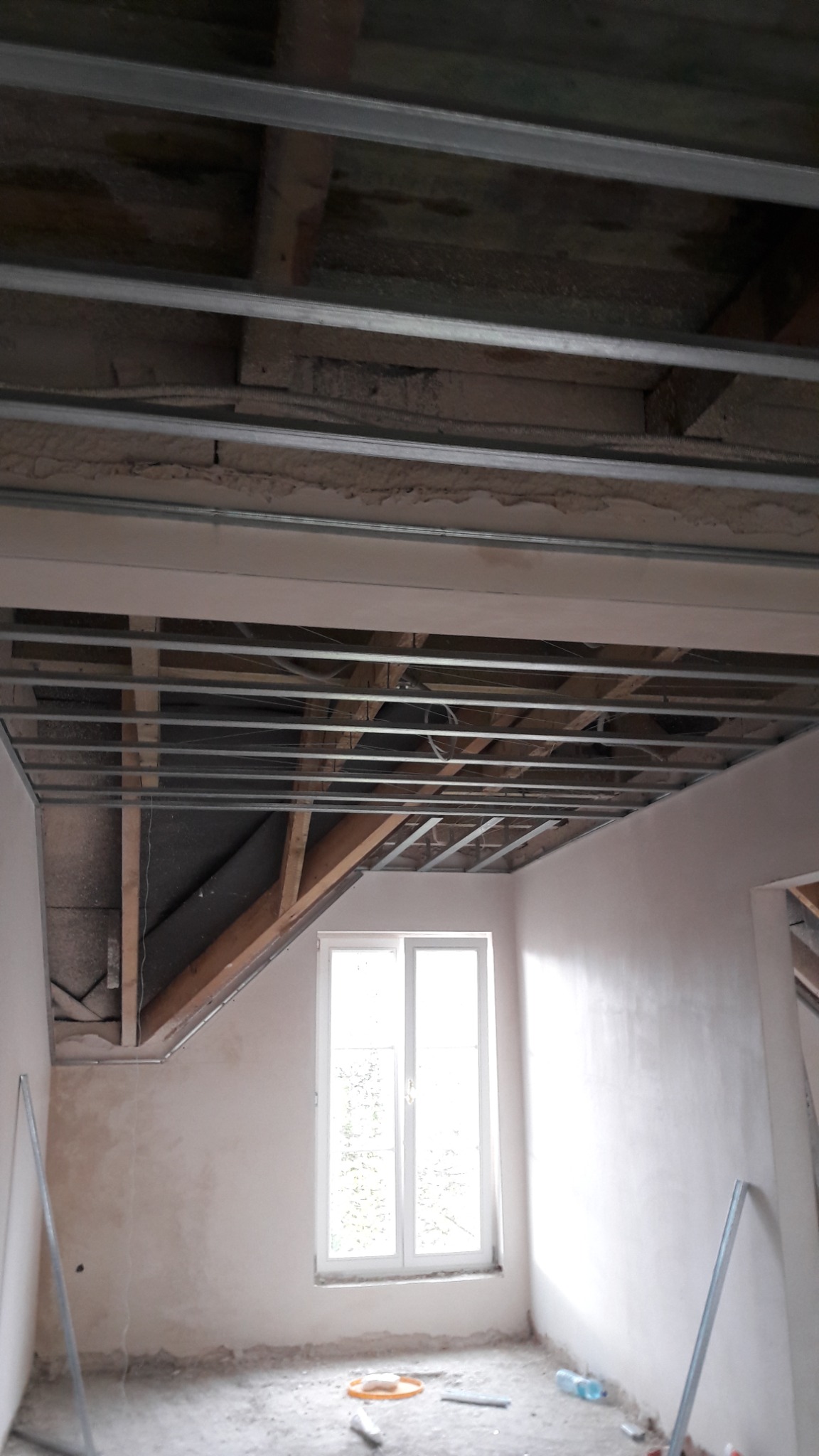 Poddasze w trakcie adaptacji: widoczne drewniane krokwie, metalowa konstrukcja sufitu podwieszanego, surowe ściany z widocznymi śladami tynku i okno w białej ramie. Na podłodze resztki materiałów...
