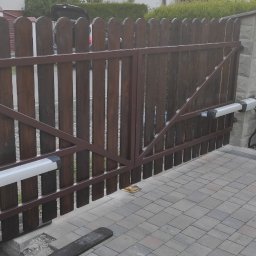 PepoAutomatyka - Drewniana brama wjazdowa z automatyką, brązowa rama, napędy ramieniowe zamontowane na słupkach, obok kwitnące róże.