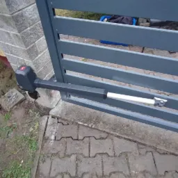 Szara brama wjazdowa z automatyką, widoczny siłownik przytwierdzony do słupka z szarej cegły, kostka brukowa na podjeździe.