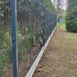 Nowoczesne ogrodzenie panelowe z podmurówką betonową, widoczne z perspektywy bocznej, z tujami w tle i słupem oświetleniowym w oddali.