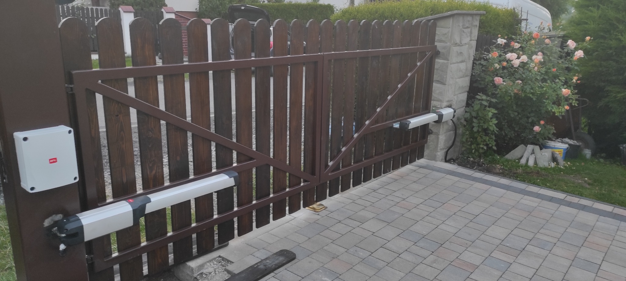Drewniana brama wjazdowa z automatyką, brązowa rama, napędy ramieniowe zamontowane na słupkach, obok kwitnące róże.