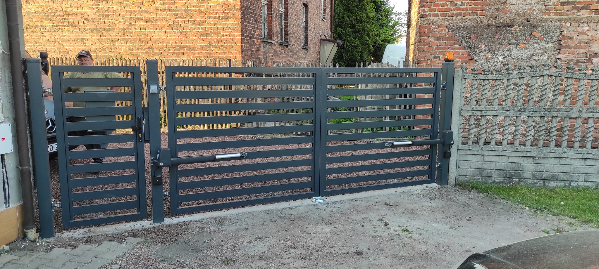 Nowoczesna, pozioma brama wjazdowa z automatyką, widoczna osoba w tle, brama w kolorze grafitowym, na posesji z ceglanym budynkiem i betonowym ogrodzeniem.