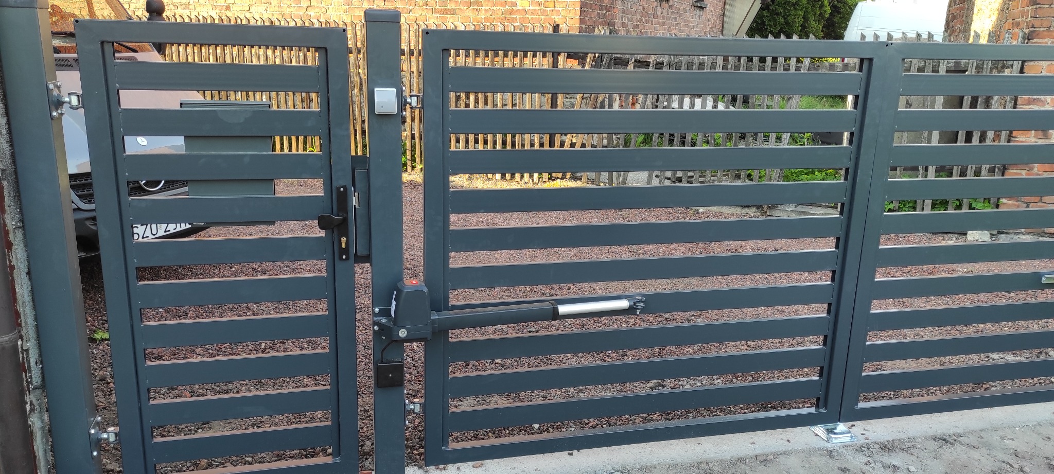 Nowoczesna, antracytowa brama wjazdowa z automatyką, widoczna w pozycji otwartej, z tłem ceglanego budynku i fragmentem zaparkowanego samochodu. Na słupku bramy zamontowany przycisk domofonu.