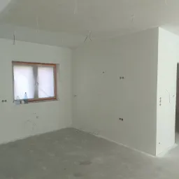 Surowe wnętrze pokoju z gładkimi, białymi ścianami, nieotynkowanym sufitem, przygotowanymi punktami elektrycznymi, oknem zabezpieczonym folią i butelkami wody na parapecie.