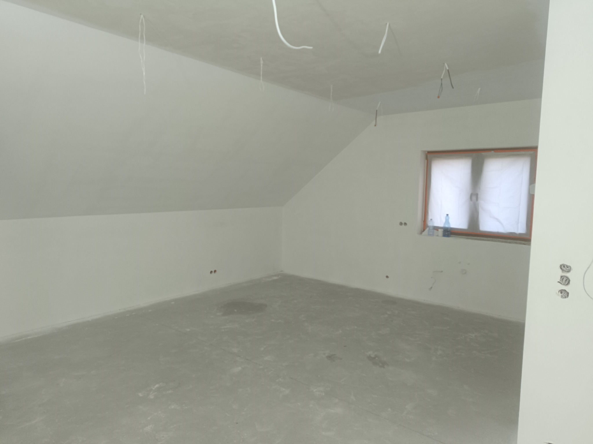 Poddasze po szpachlowaniu, ściany i sufit na biało, widoczne wystające przewody elektryczne i okno zabezpieczone folią malarską.