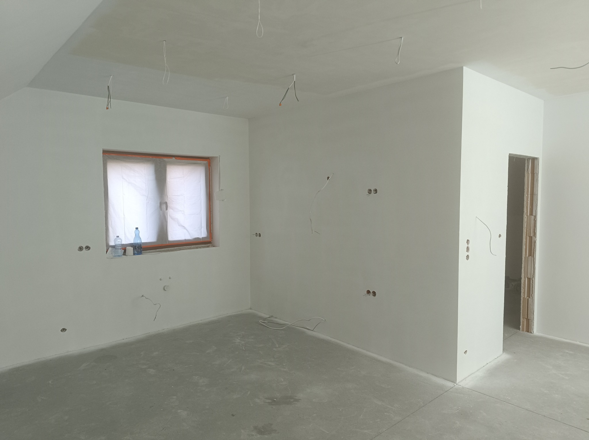 Surowe wnętrze pokoju z gładkimi, białymi ścianami, nieotynkowanym sufitem, przygotowanymi punktami elektrycznymi, oknem zabezpieczonym folią i butelkami wody na parapecie.