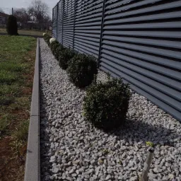 Nowoczesny, szary płot lamelowy z nasadzeniami z bukszpanu i białych kulistych lamp, podsypane drobnym, szarym kamieniem.