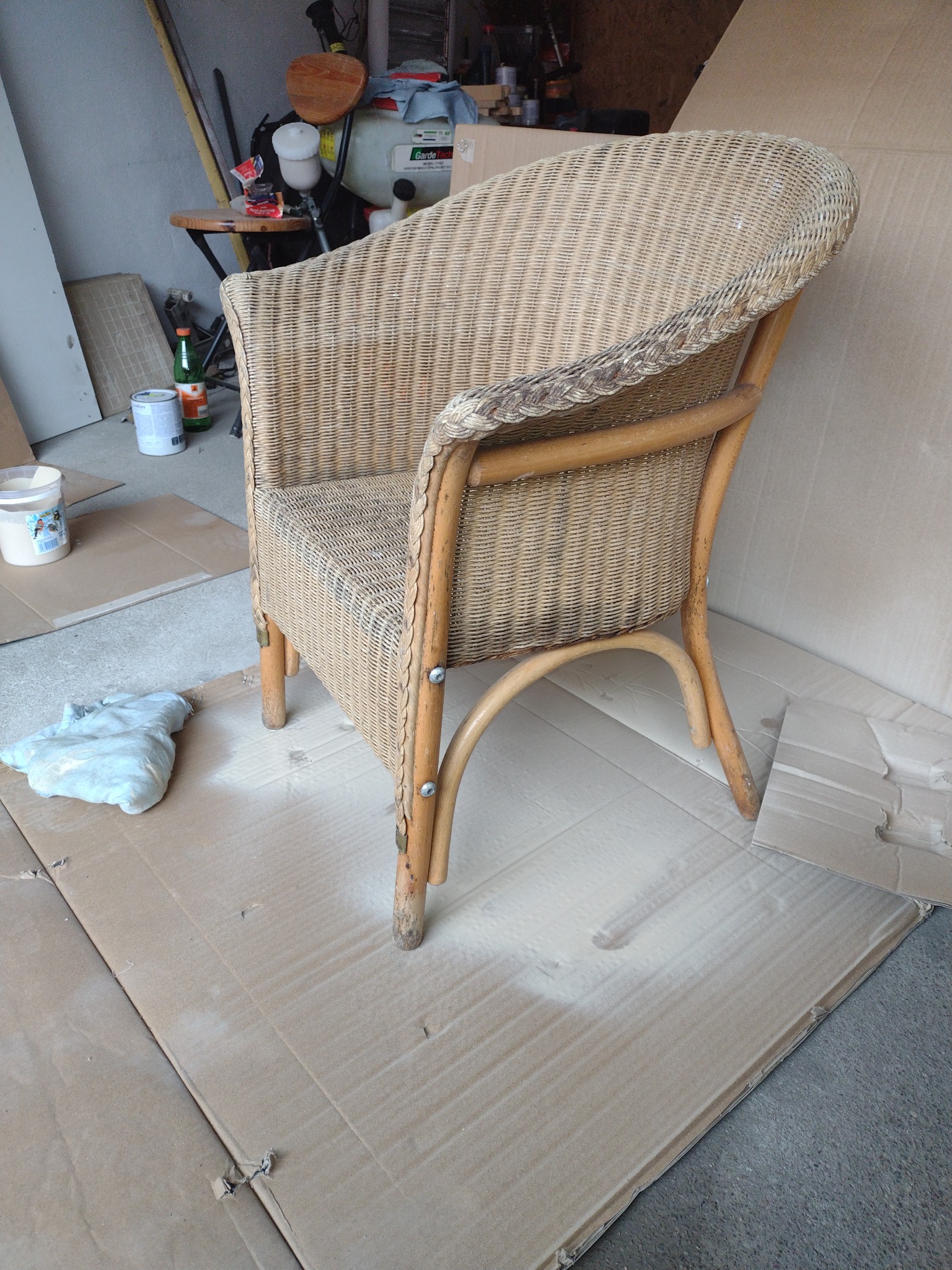 Rattanowy fotel przygotowany do malowania, zabezpieczony kartonem, widoczny pojemnik z farbą i szmatka.