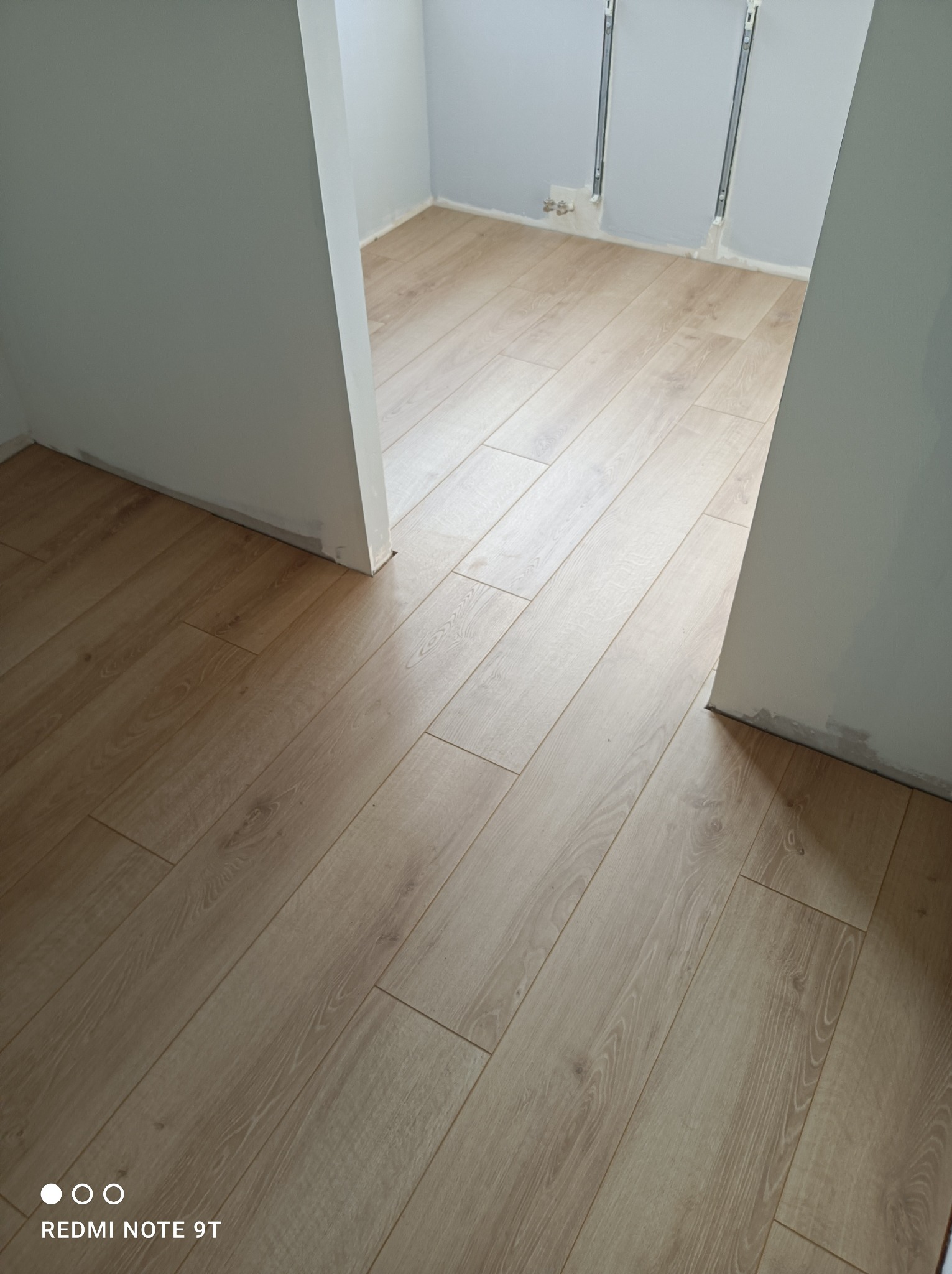 Jasne panele podłogowe ułożone w pustym pokoju z szarymi ścianami, widoczne ślady po malowaniu na ścianach przy podłodze.