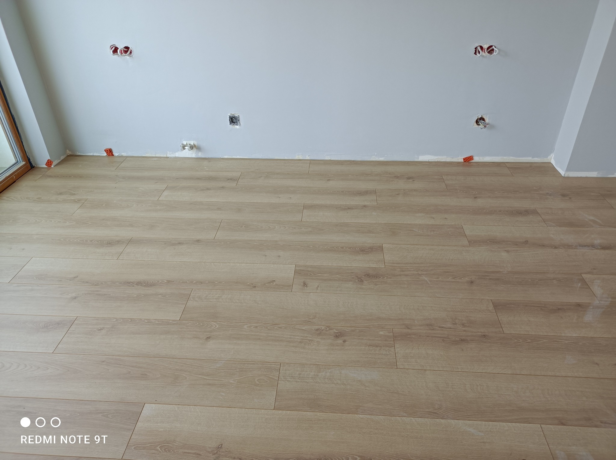 Jasne panele podłogowe ułożone w pokoju z szarą ścianą i wystającymi kablami elektrycznymi, widoczne pomarańczowe kliny dystansowe przy ścianie.