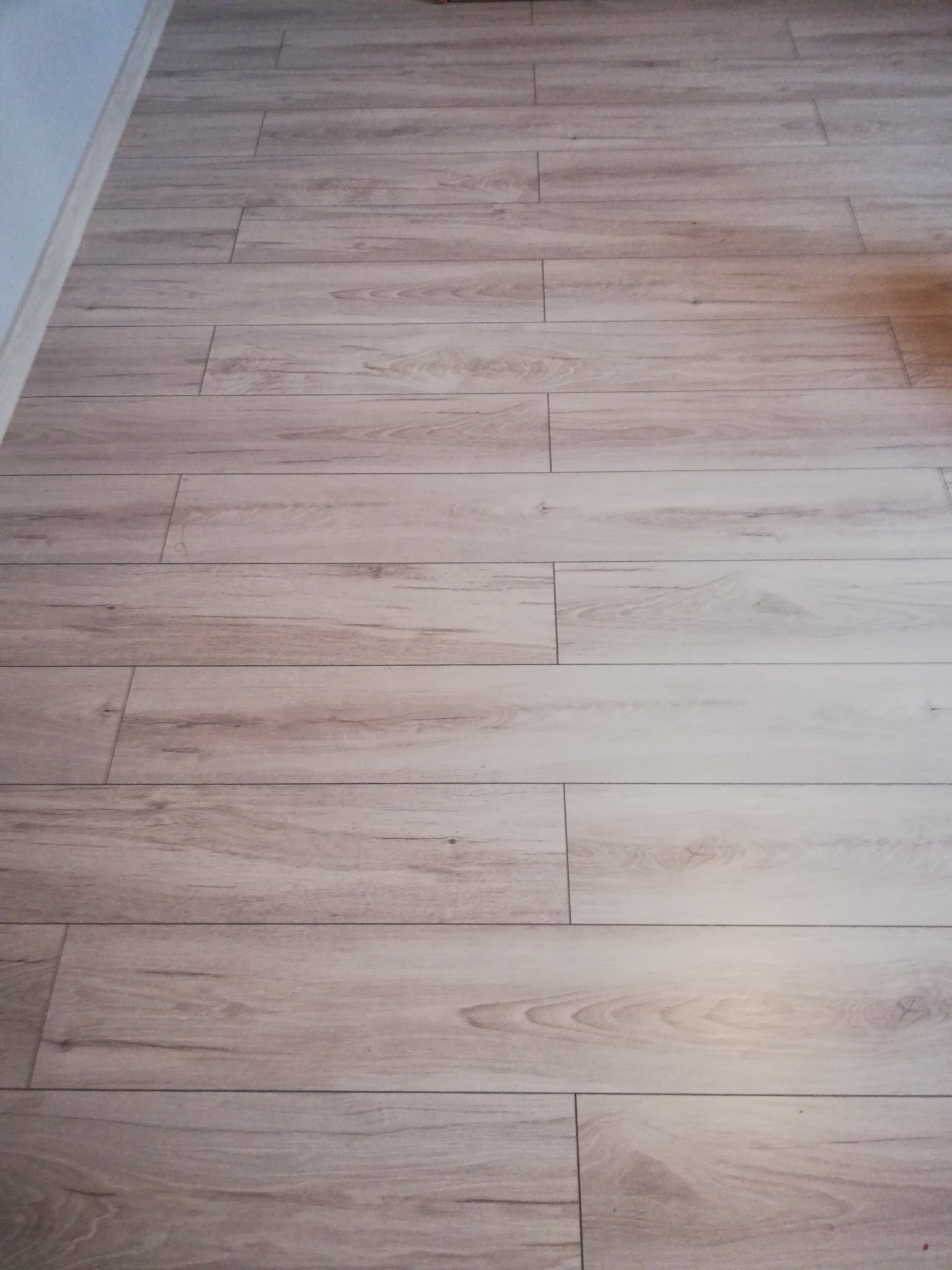 Jasne panele podłogowe ułożone wzdłuż pomieszczenia, widoczna ściana w tle po lewej stronie.