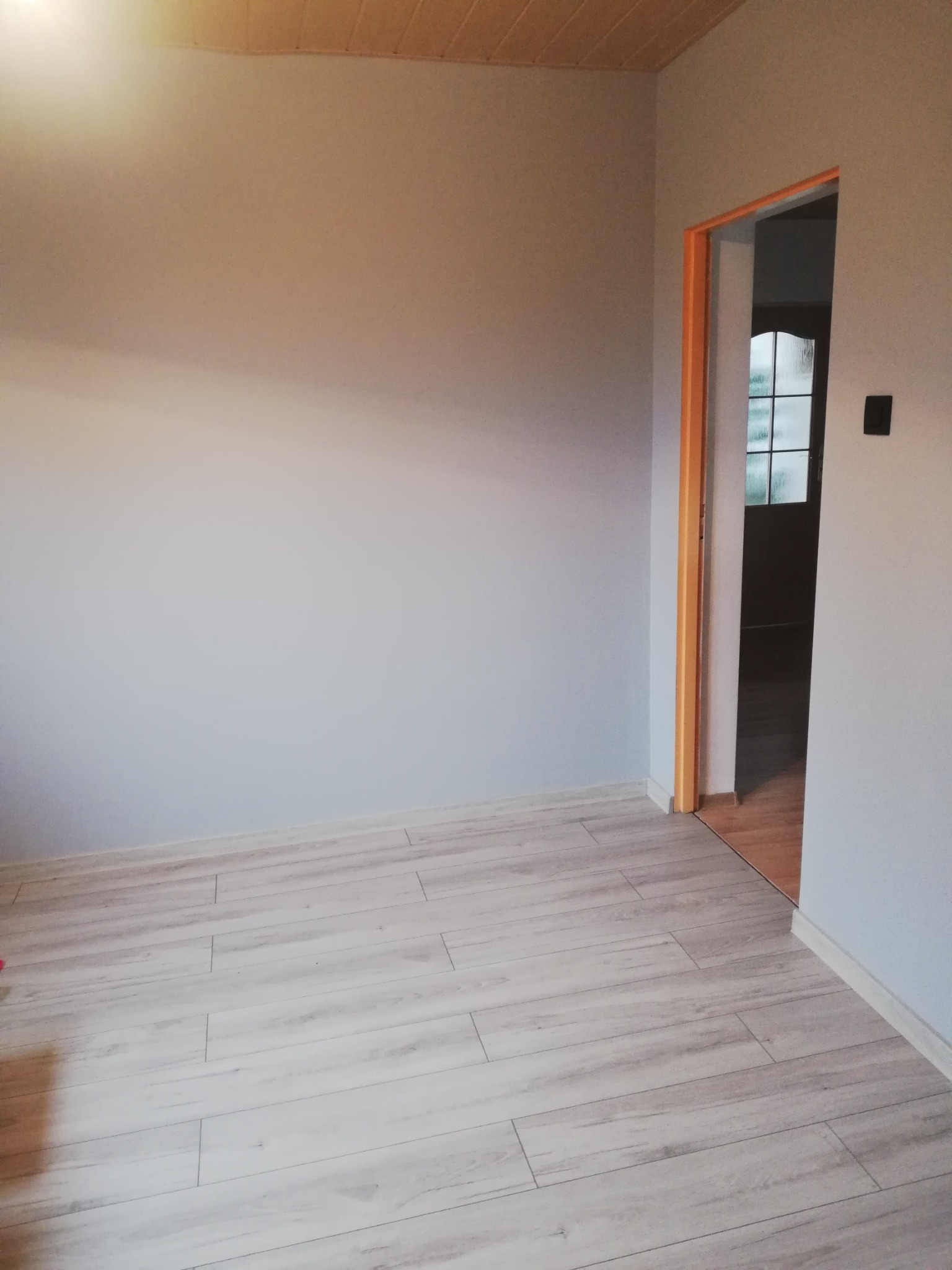 Pomalowane na szaro puste pomieszczenie z jasnymi panelami podłogowymi imitującymi drewno i drewnianą boazerią na suficie, widok na sąsiedni pokój przez otwarte drzwi z pomarańczową futryną.