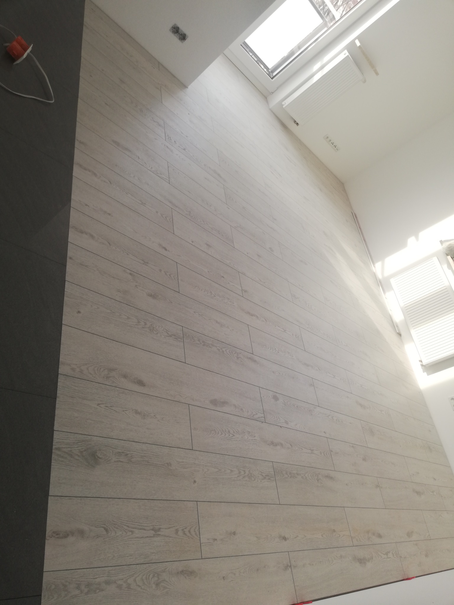 Ściana wyłożona jasnymi panelami podłogowymi z widocznym wzorem drewna, obok ciemnoszara ściana, okno dachowe i grzejnik w jasnym pomieszczeniu.