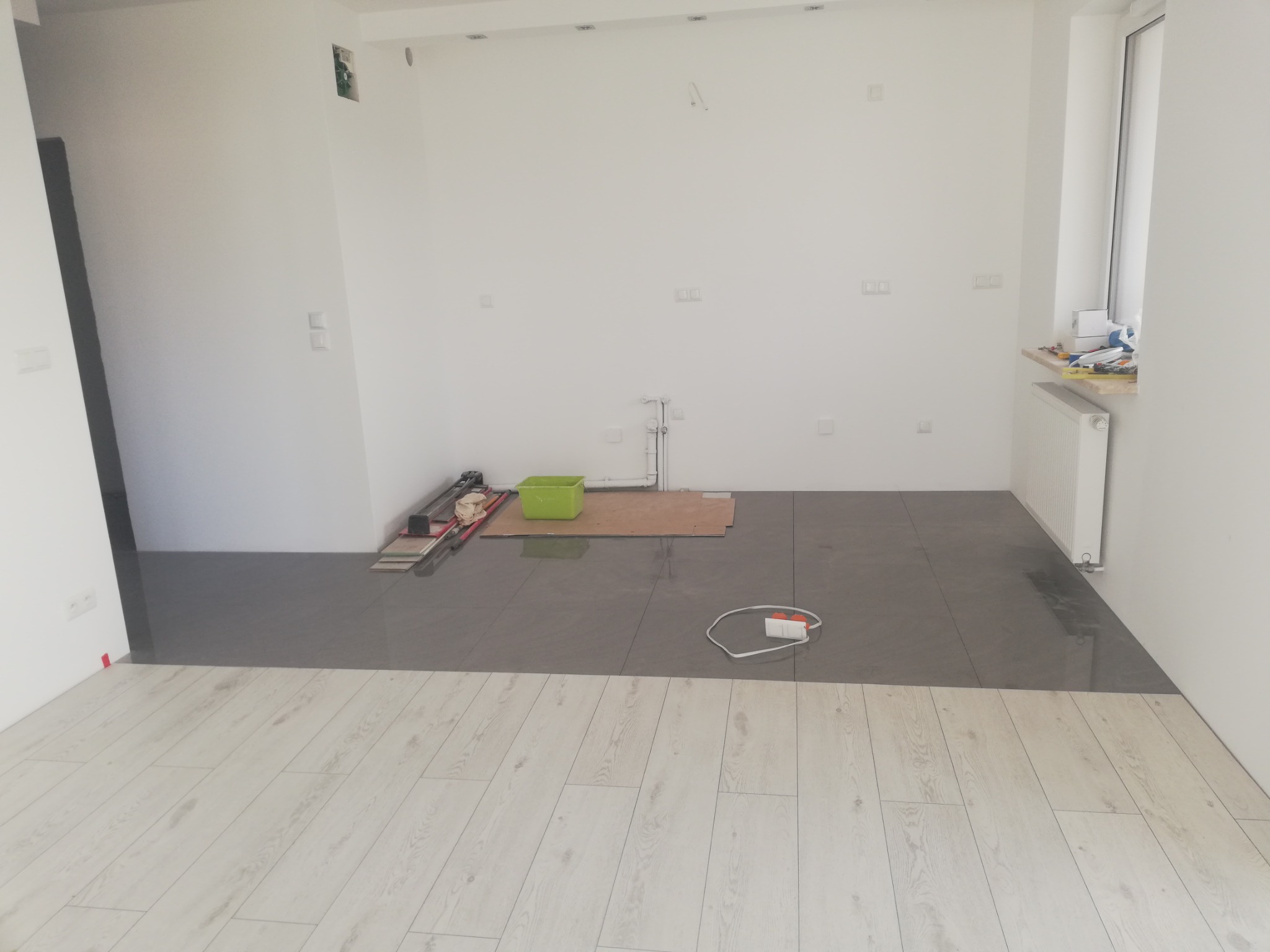 Wnętrze w trakcie remontu, z fragmentem ułożonych szarych płytek i jasnych paneli podłogowych, widoczne narzędzia i akcesoria budowlane, ściany w trakcie przygotowania pod malowanie.