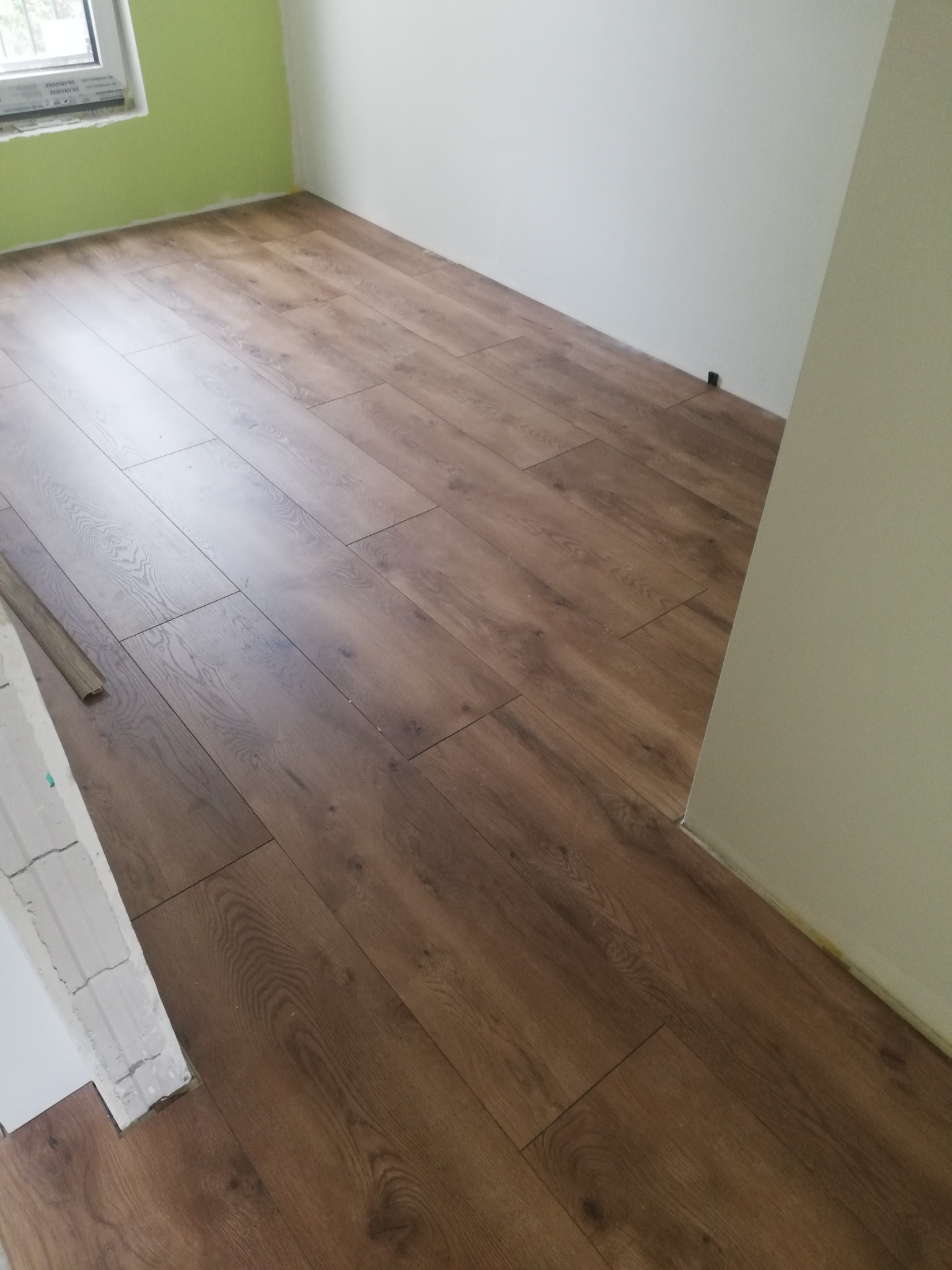 Ułożone panele podłogowe w odcieniach brązu w rogu pokoju, widoczne niedokończone prace przy listwie przypodłogowej i fragment zielonej ściany.