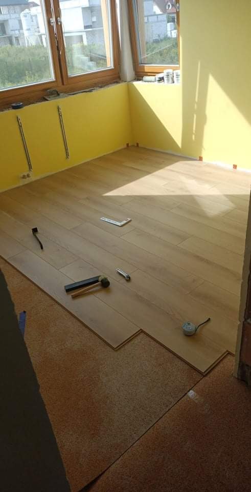 Fragment pokoju z żółtymi ścianami, w trakcie montażu jasnych paneli podłogowych na podkładzie korkowym, widoczne narzędzia: młotek gumowy, miarka, kątownik.