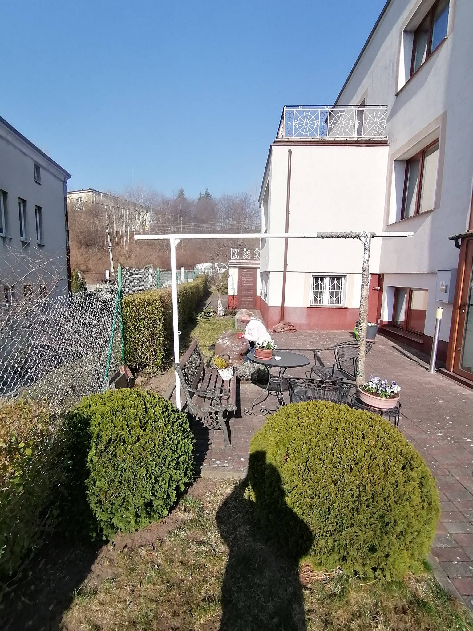 Ogród z dwoma kulistymi krzewami na pierwszym planie, metalowy zestaw mebli ogrodowych na tarasie z cegły, kobieta przy stole, budynki w tle, cień osoby robiącej zdjęcie.