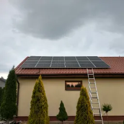 Panele fotowoltaiczne zamontowane na dachu budynku z czerwoną dachówką, drabina oparta o dach, pochmurne niebo w tle.